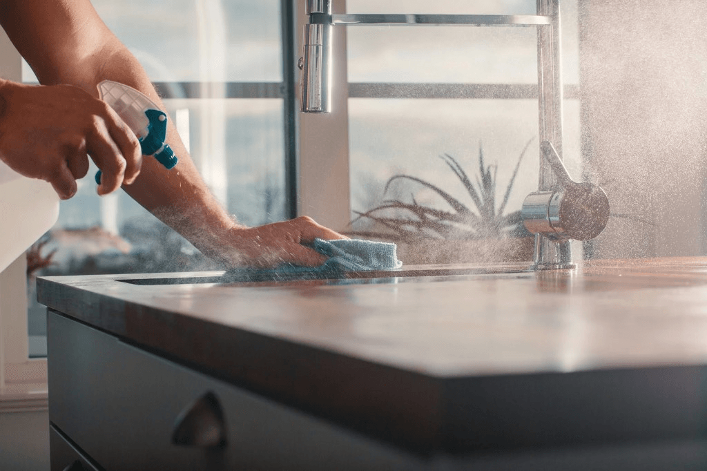 Hands spraying and wiping a kitchen countertop with a microfiber cloth in a sunlit kitchen